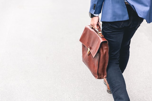 A Man In A Business Suit, Walking With His Leather Bag In Hand.